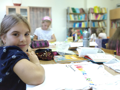 Dziewczynka na pierwszym planie siedząca przy stoliku. W tle jej koleżanki projektujące koszulki.
