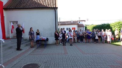 Zdjęcie dziedzińca szkolnego podczas uroczystości. Osoby prowadzące, dyrektor szkoły oraz poczet flagowy.