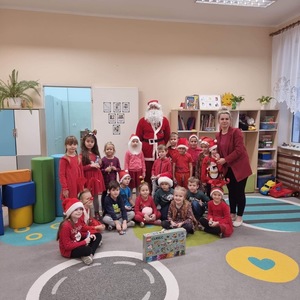 Święty Mikołaj z grupą przedszkolną dzieci ubranych w czerwone stroje. Przed nimi stoi zestaw klocków LEGO.