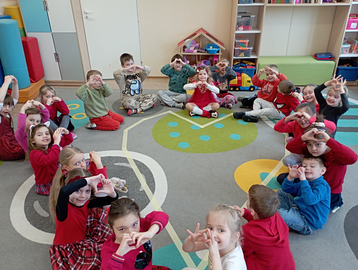 Dzieci tworzą kształt serca siedząc na dywanie, pokazując dłońmi nad głowami kształt serca.