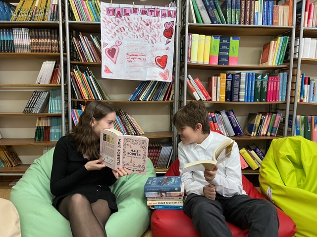 Uczennica i uczeń kl. 8 siedzą na pufach w bibliotece  z książkami w ręku i rozmawiają ze sobą.