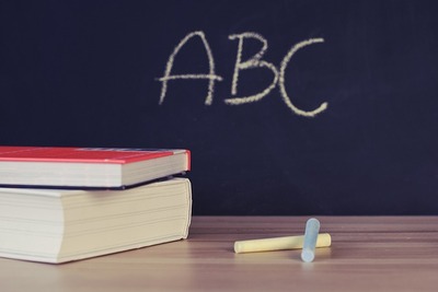 Zdjęcie przedstawiające książki i kredę leżące na biurku na tle tablicy szkolnej. Obraz StockSnap z Pixabay
