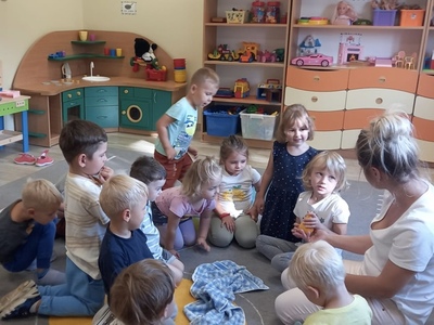 Dzieci siedzą w kole wraz z nauczycielem, dziewczynka trzyma w ręku żółta plastikową gruszkę, którą wyjęła spod materiału, który znajduje się w środku koła.