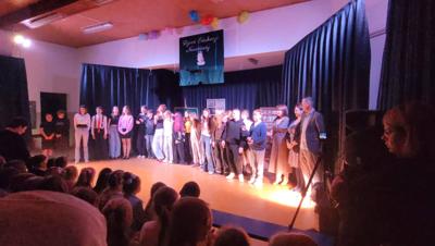 Widok sceny z grupą aktorów i opiekunów.