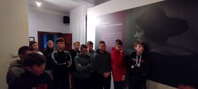 Grupa uczniów w sali muzealnej słucha opowiadania przewodnika.