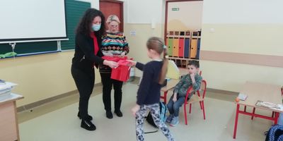 Dziewczynka odbiera walentynkę. Przed nią nauczycielka w maseczce 