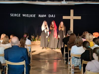 Scena z krzyżem, na sali ludzie siedzący na krzesłach. Na scenie trzy dziewczynki Przedstawienie Wielkanocne 13.04.2022 r.