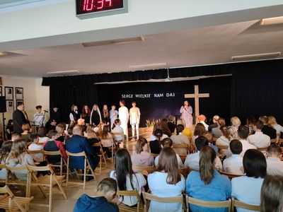 Scena z krzyżem, ludzie siedzący na krzesłach. Na scenie kilka osób które uczestniczyły  w przedstaieniu.