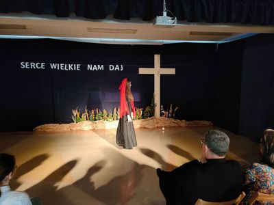 Widok sceny z krzyżem oraz kobiety w czarnej sukni i czerwonej chuście
