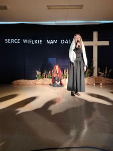 Scena z dwoma kobietami w czarnych sukniach, pierwsza trzyma mikrofon, druga siedzi okryta czerwoną chustą.