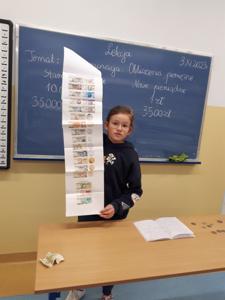 Uczennica trzymająca listę banknotów sprzed denominacji w porównaniu do dzisiejszych kwot i monet.