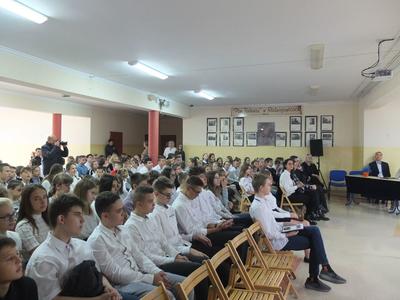 Uczniowie siedzący na krzesłach w białych koszulach.