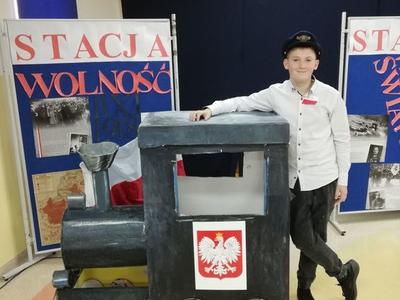 Chłopiec z biało czerwonym kotylionem stoi obok makiety pociągu.