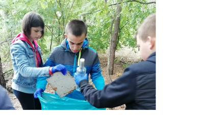 Dwóch uczniów i uczennica zbierają butelki do niebieskiego worka.