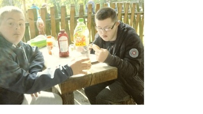 Dwóch uczniów siedzi przy drewnianym stoliku i jedzą upieczone na ognisku kiełbaski.