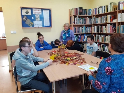 Pani bibliotekarka czyta uczniom, którzy siedzą wokół stołu wiersze o tematyce jesiennej.
