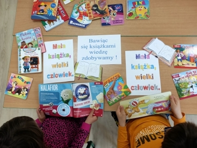 Madzia i Piotruś oglądają kolorowe książki. Na stole porozkładane plakaty z hasłami „Mała książka, wielki człowiek”, „Bawiąc się książkami, wiedzę zdobywamy”.