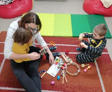 Pani Agnieszka i dzieci siedzą na dywanie. Obok rozsypane instrumenty muzyczne.