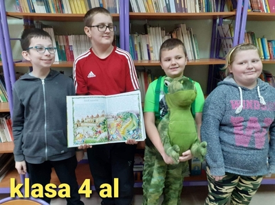 Na zdjęciu klasa 4 al. W bibliotece szkolnej. Jeden z uczniów trzyma książkę a drugi maskotkę.Cała klasa jest uśmiechnięta.
