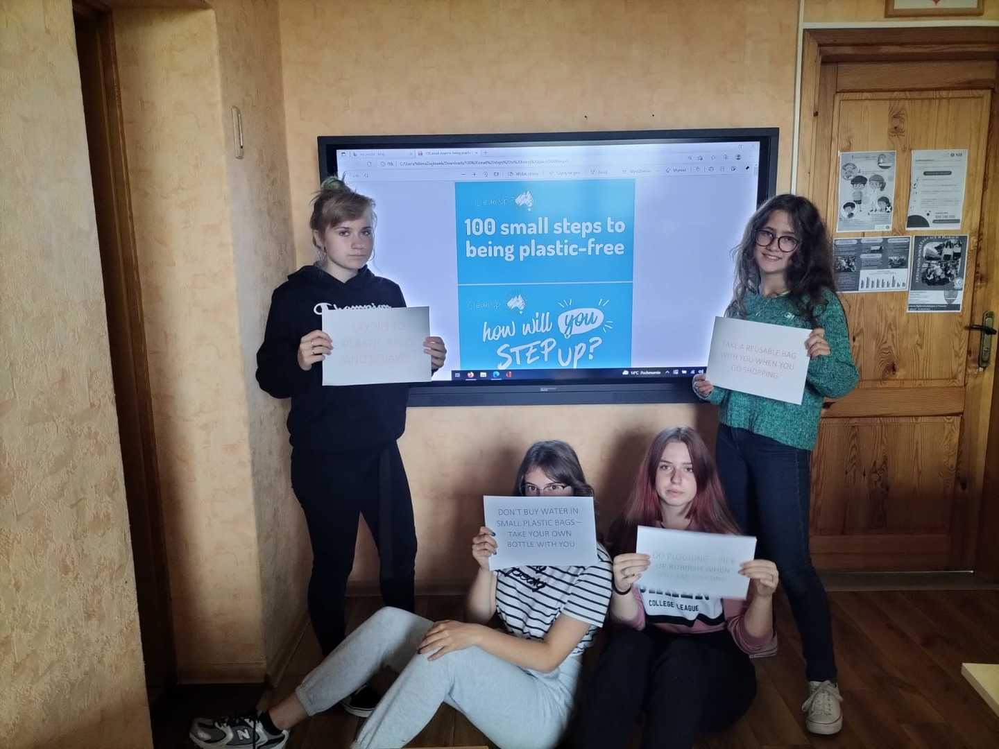 Cztery uczennice z klasy VIII prezentują hasła ekologiczne napisane w języku angielskim. W tle monitor z hasłem przewodnim.