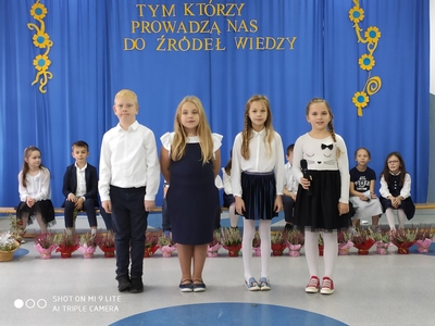 Uczniowie odświętnie ubrani, stoją na środku sali i z okazji święta Dnia Edukacji Narodowej i recytują wiersze. W tle niebieska kotara z hasłem uroczystości, a po bokach ozdobne dekoracje z kwiatków. Przed kurtyną, na ławce, siedzą uczniowie z klasy II. Przed uczniami, na podłodze, ustawione są kolorowe wrzosy.