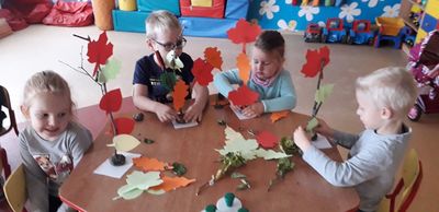 Dwie dziewczynki i dwóch chłopców siedzą przy stoliku w przedszkolu i wykonują prace z jesiennych liści.