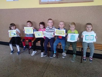 Uśmiechnięte dzieci siedzą na ławce prezentując kartki z wesołymi wierszykami.