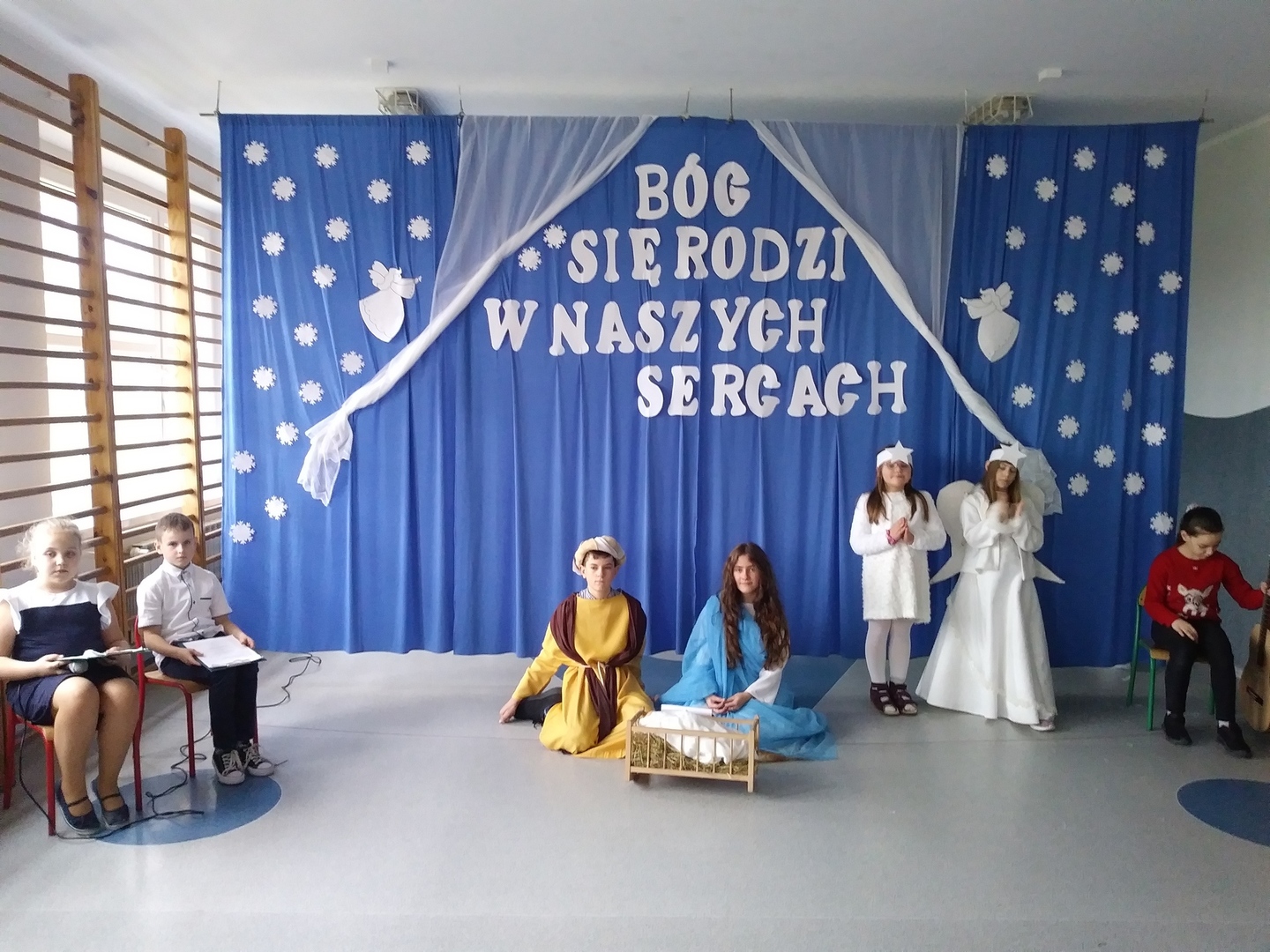 W tle niebieskiej dekoracji z napisem „Bóg się rodzi w naszych sercach” uczniowie odgrywający role w jasełkach.