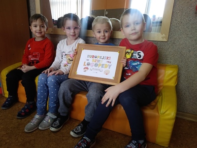 Uśmiechnięci przedszkolacy, jeden z nich trzyma tabliczkę z napisem Europejski Dzień Logopedy.