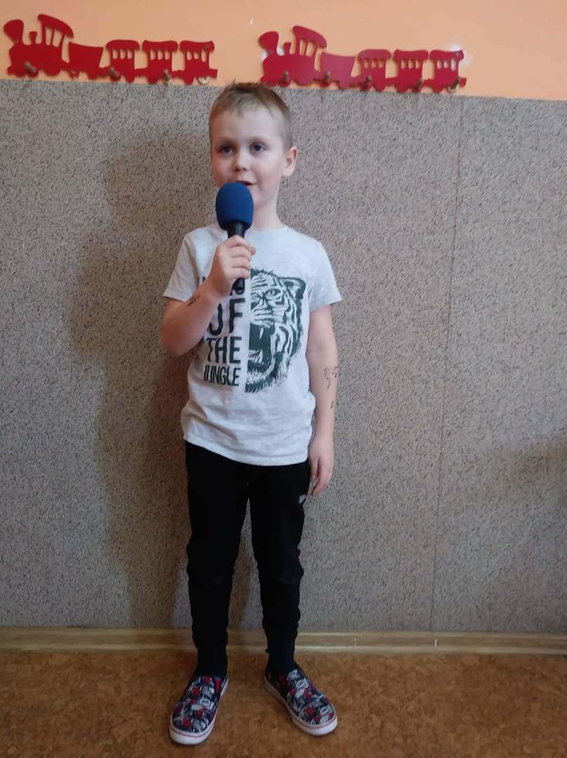 Na pierwszym planie chłopczyk z mikrofonem recytujący wierszyk.