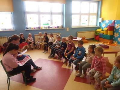 W sali przedszkolnej uczennice klasy V oraz grupa przedszkolna słuchająca czytanej im bajeczki.