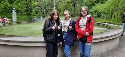Trzy uczennice z naszej szkoły biorące udział w wycieczce: Wiktoria Łapok z klasy 8 oraz Julia Kot i Nina Osadnik z klasy 7. Za nimi fontanna i zieleń w parku. Na ławkach siedzą przypadkowi zwiedzający.