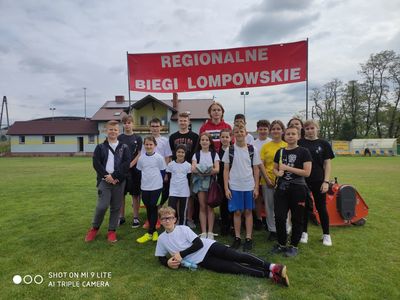 Dziewczęta i chłopcy reprezentujący naszą szkołę na zawodach sportowych na murawie boiska sportowego. Za uczniami transparent informujący o nazwie zawodów. 