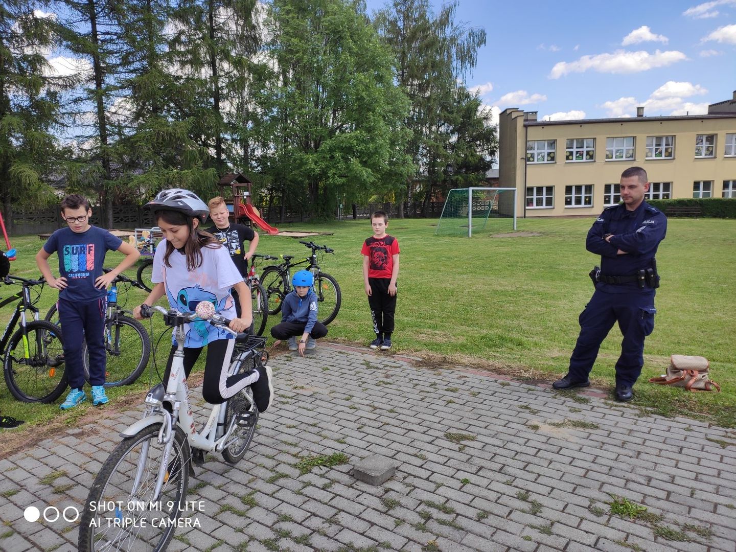 Policjant nadzorujący przebieg egzaminu na kartę rowerową. Z lewej strony uczniowie ze swoimi rowerami. Na pierwszym planie uczennica na rowerze w kasku na głowie w trakcie pokonywania toru przeszkód. W tle trawiaste boisko szkolne oraz budynek szkoły.