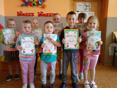 Na zdjęciu uśmiechnięci przedszkolacy, którzy uczęszczali na zajęcia logopedyczne. Dzieci prezentują otrzymane dyplomy za udział w zajęciach. W tle sala lekcyjna.