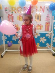 Zdjęcie przedstawia uśmiechniętą Michalinkę z balonikiem, w tle tablica z napisem Urodziny.