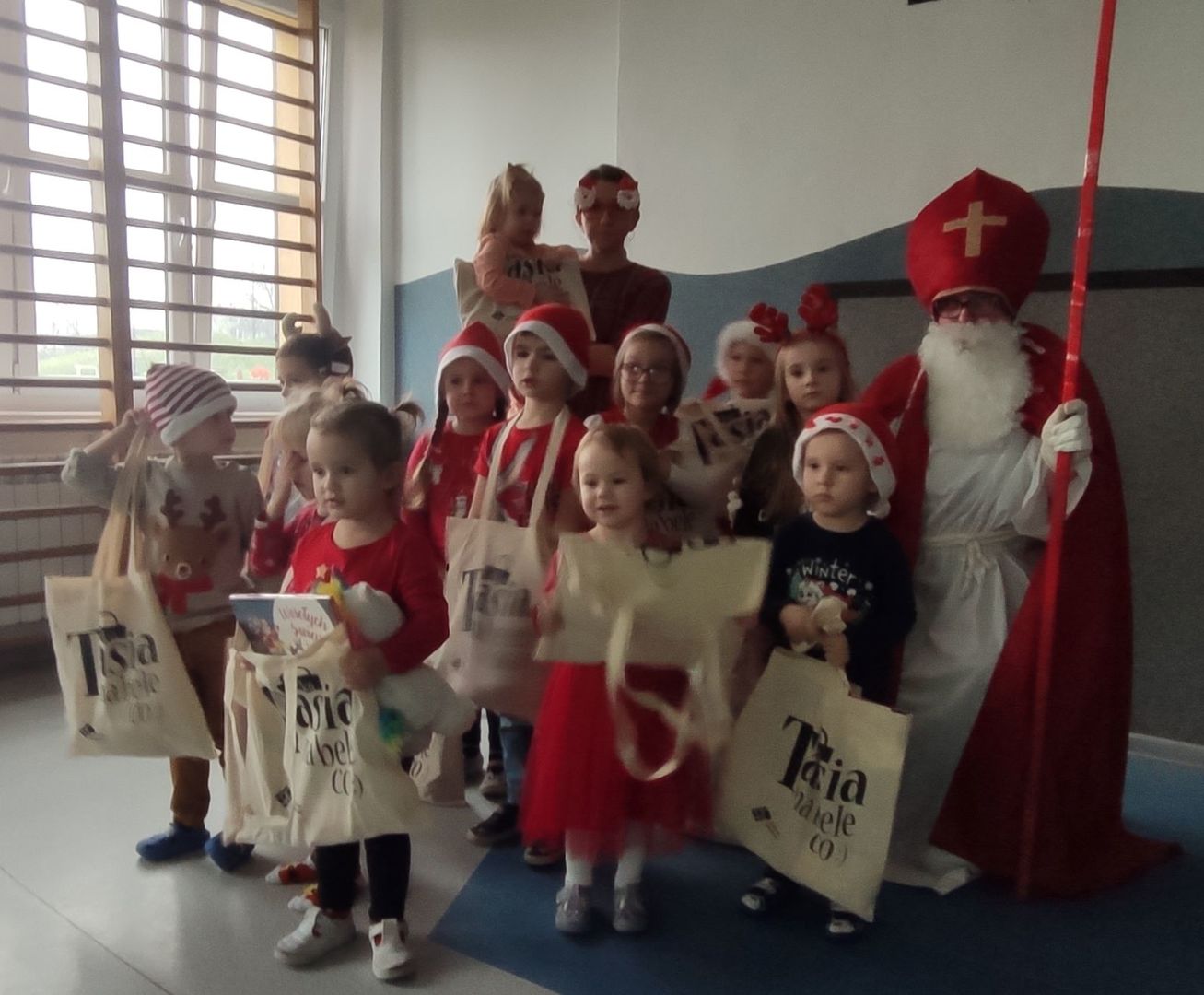 Dzieci z przedszkola w czerwonych czapkach z prezentami, Świętym Mikołajem i panią przedszkolanką.