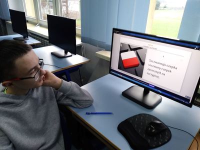 Zdjęcie przedstawia ucznia klasy VI podczas ćwiczeń z komputerem, w tle pracownia komputerowa.