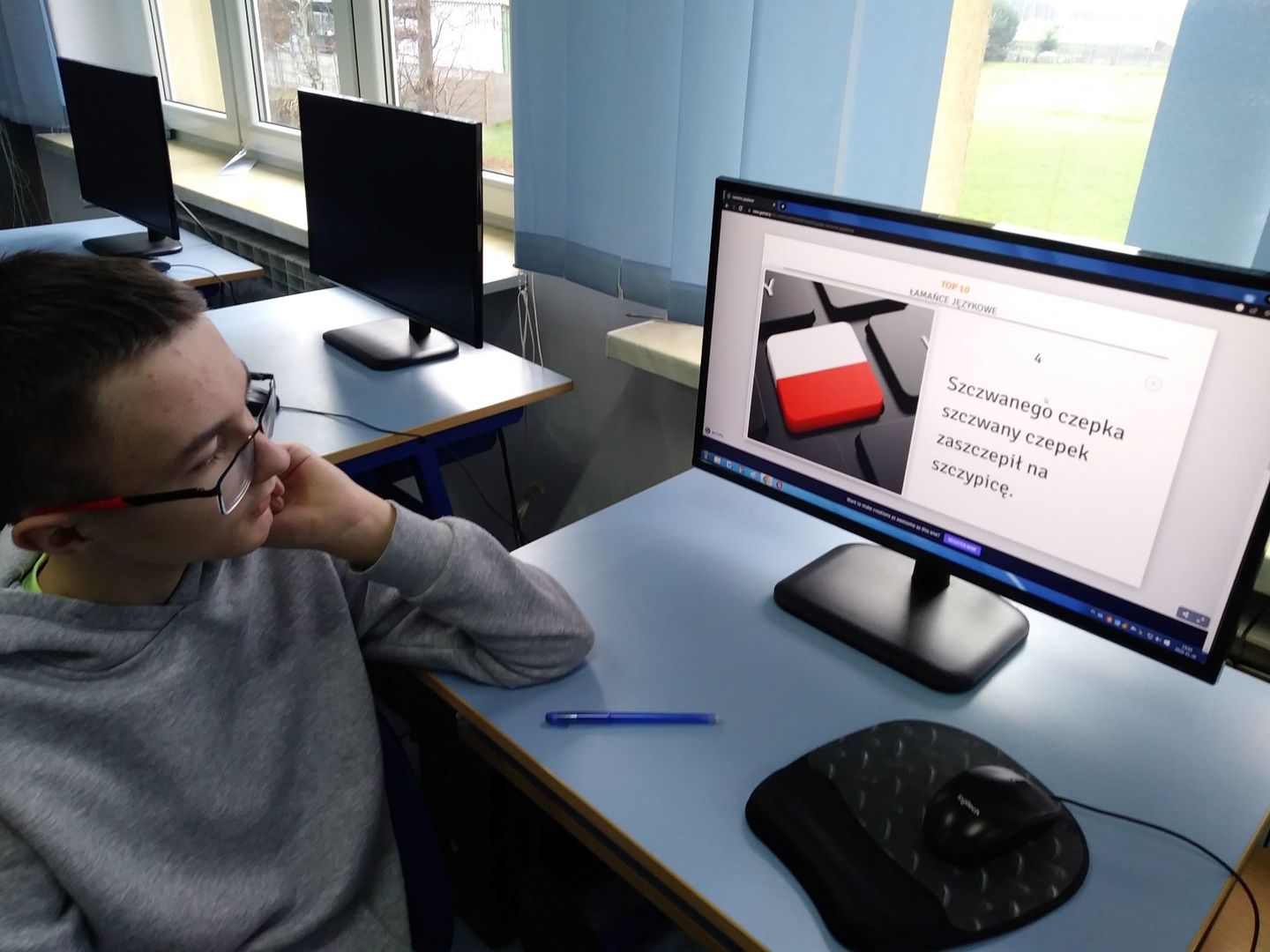 Zdjęcie przedstawia ucznia klasy VI podczas ćwiczeń z komputerem, w tle pracownia komputerowa.