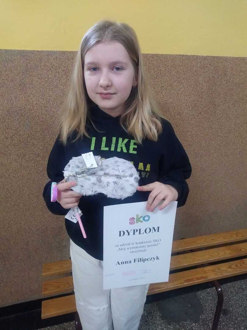 Zdjęcie przedstawia uczennicę klasy V Anię wraz z dyplomem oraz portfelem, który samodzielnie wykonała, w tle korytarz szkolny.
