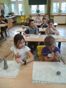 Uczniowie klasy 2b, siedzący przy stolikach podczas wykonywania skrzatów z gliny.