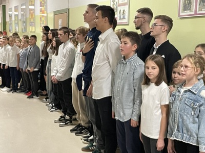 Grupa uczniów w strojach galowych w korytarzu szkolnym śpiewa hymn narodowy