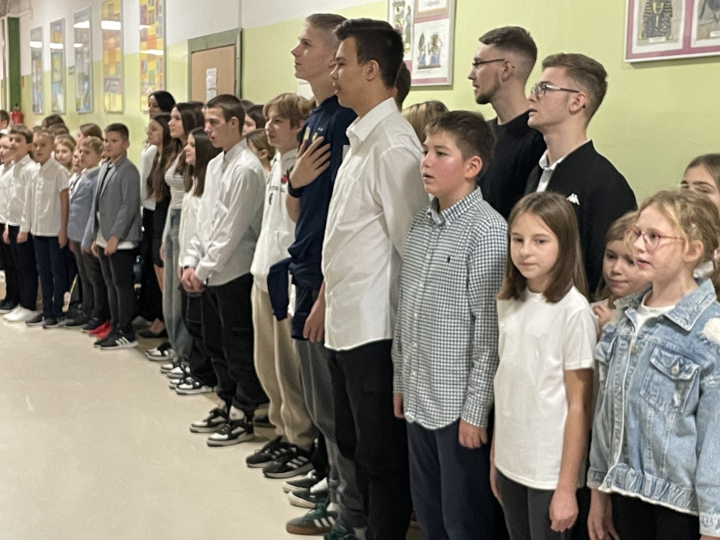 Grupa uczniów w strojach galowych w korytarzu szkolnym śpiewa hymn narodowy