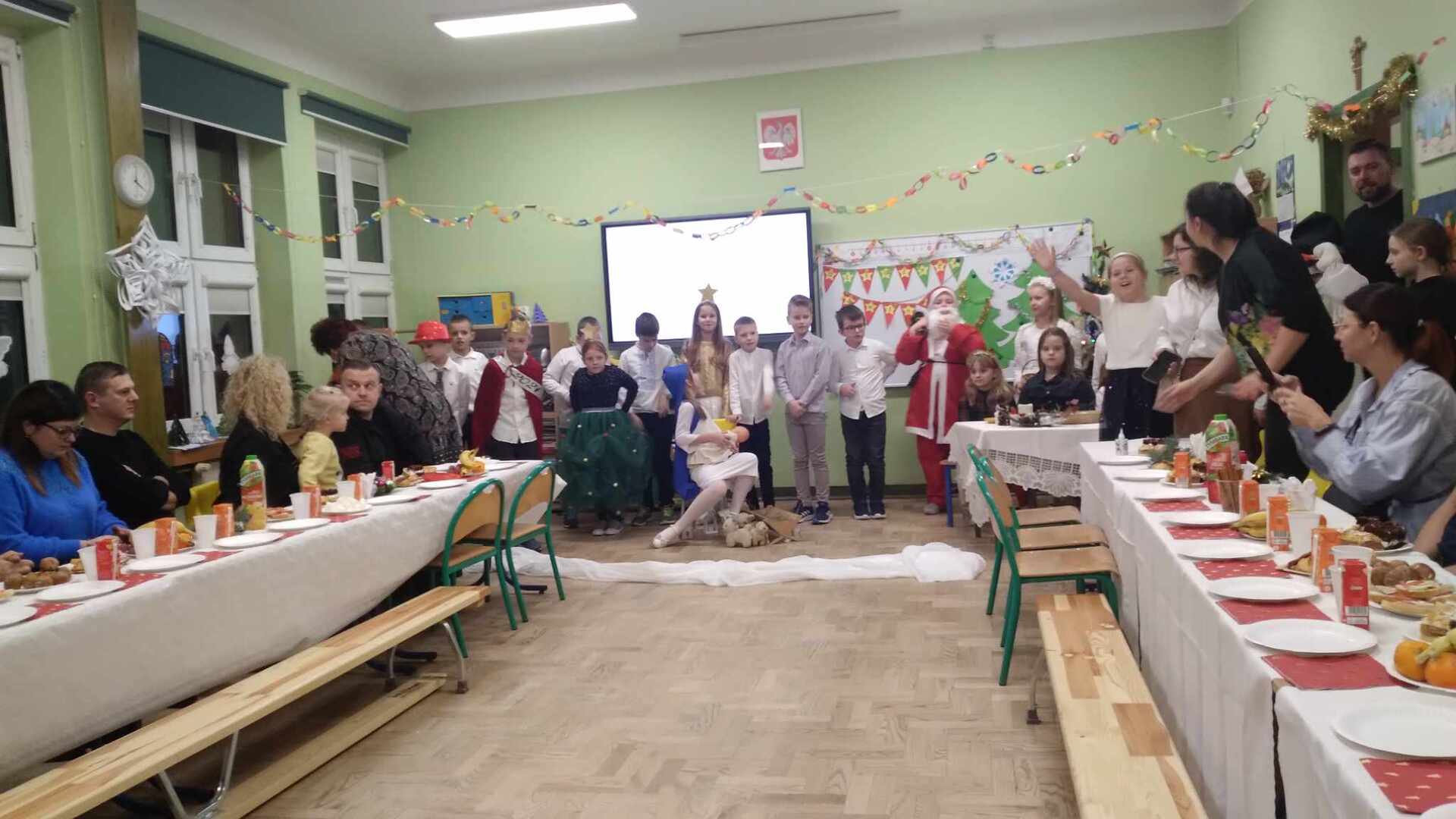 rodzice uczniów, siedzący za stołami w sali lekcyjnej, oglądają występy dzieci