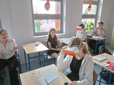 Uczniowie w sali lekcyjnej siedzą w ławkach, mają na sobie okulary VR