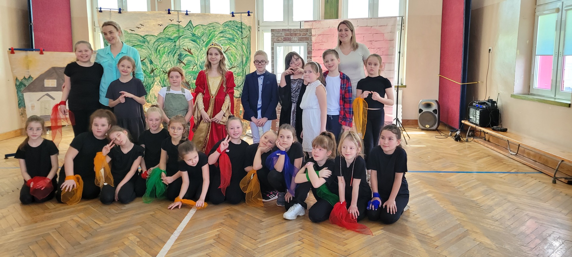 Grupa dzieci pozuje do zdjęcia po przedstawieniu na scenie, przed dekoracją lasu.