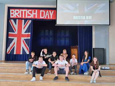 Grupa uczniów siedzi na schodkach auli 26. L.O. w Łodzi. W tle flaga brytyjska i napis British Day.
