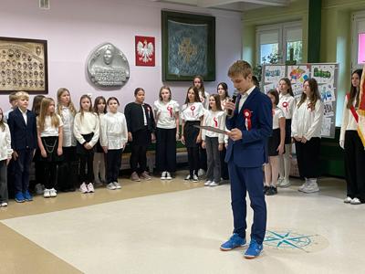 Uczniowie w strojach galowych stoją w holu szkoły. jeden z uczniów czyta informacje z kartki.