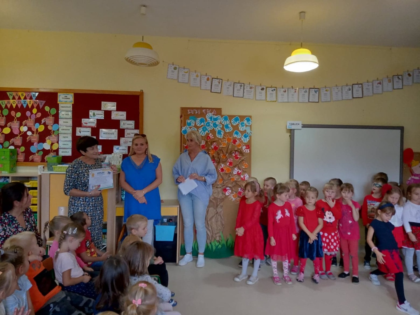 Dyrektor wręcza pani burmistrz dyplom, dzieci w czerwonych strojach patrzą z zaciekawieniem