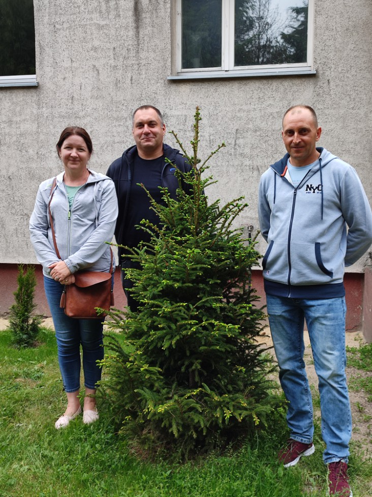 Zdjęcie przedstawia troje rodziców (od lewej: jedna kobiet i dwóch mężczyzn) stojących za niedużym drzewkiem iglastym.